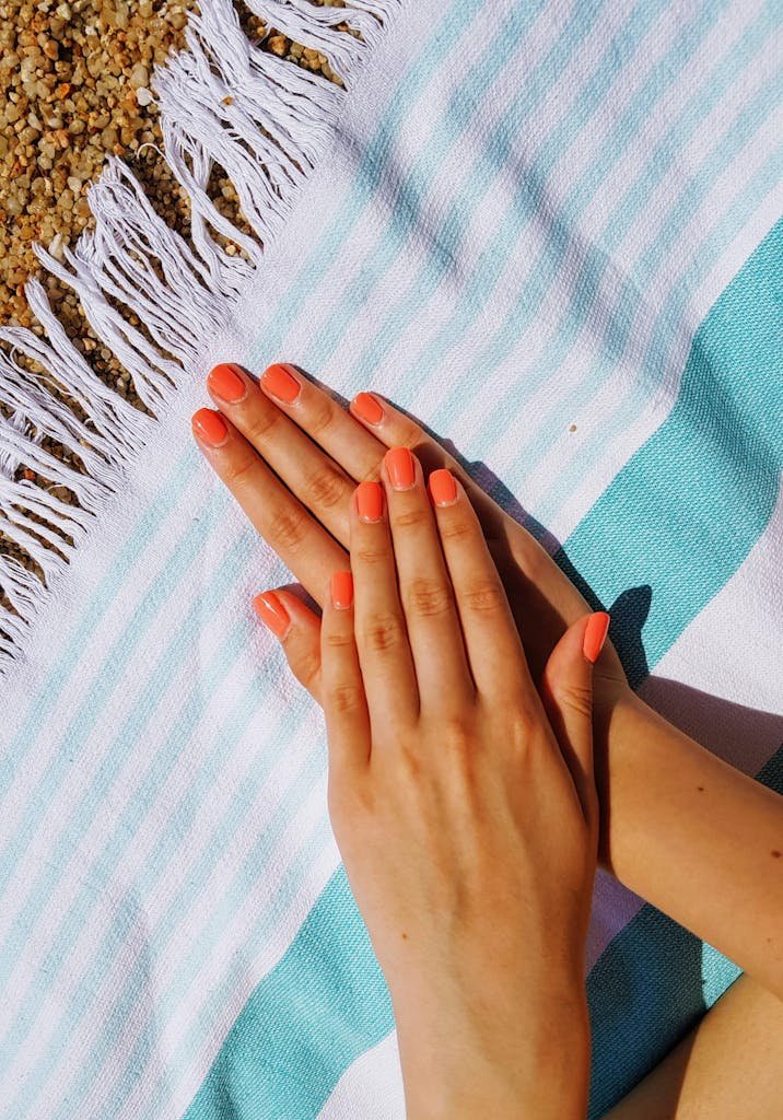 Two hands with coral nail polish resting on a striped beach towel, capturing a summer vibe.