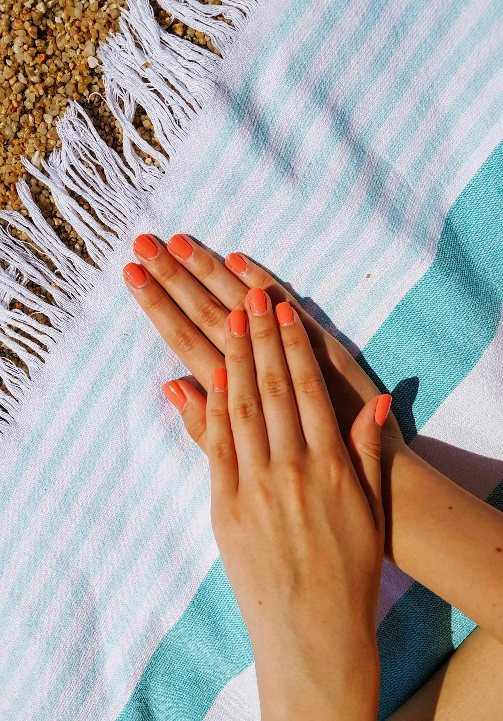 Two hands with coral nail polish resting on a striped beach towel, capturing a summer vibe.